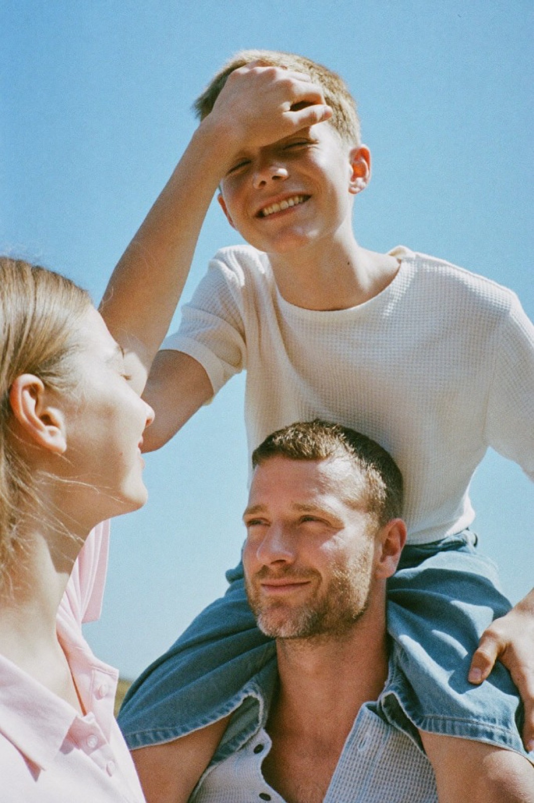 A boy on his father's shoulders under a blue sky, shading his eyes and laughing