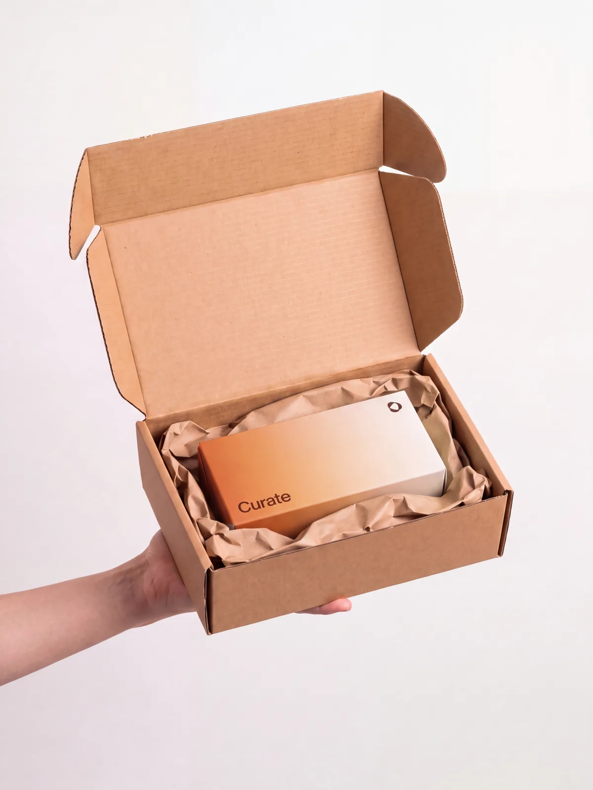 A hand holding an open, unmarked cardboard delivery box with the orange Curate carton inside on kraft paper