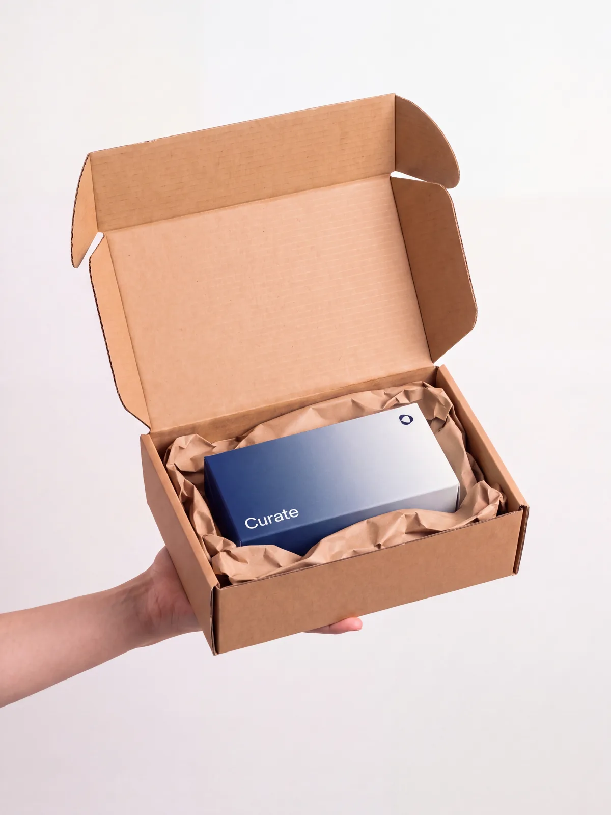 A hand holding an open, unmarked cardboard delivery box with the navy Curate carton inside on kraft paper