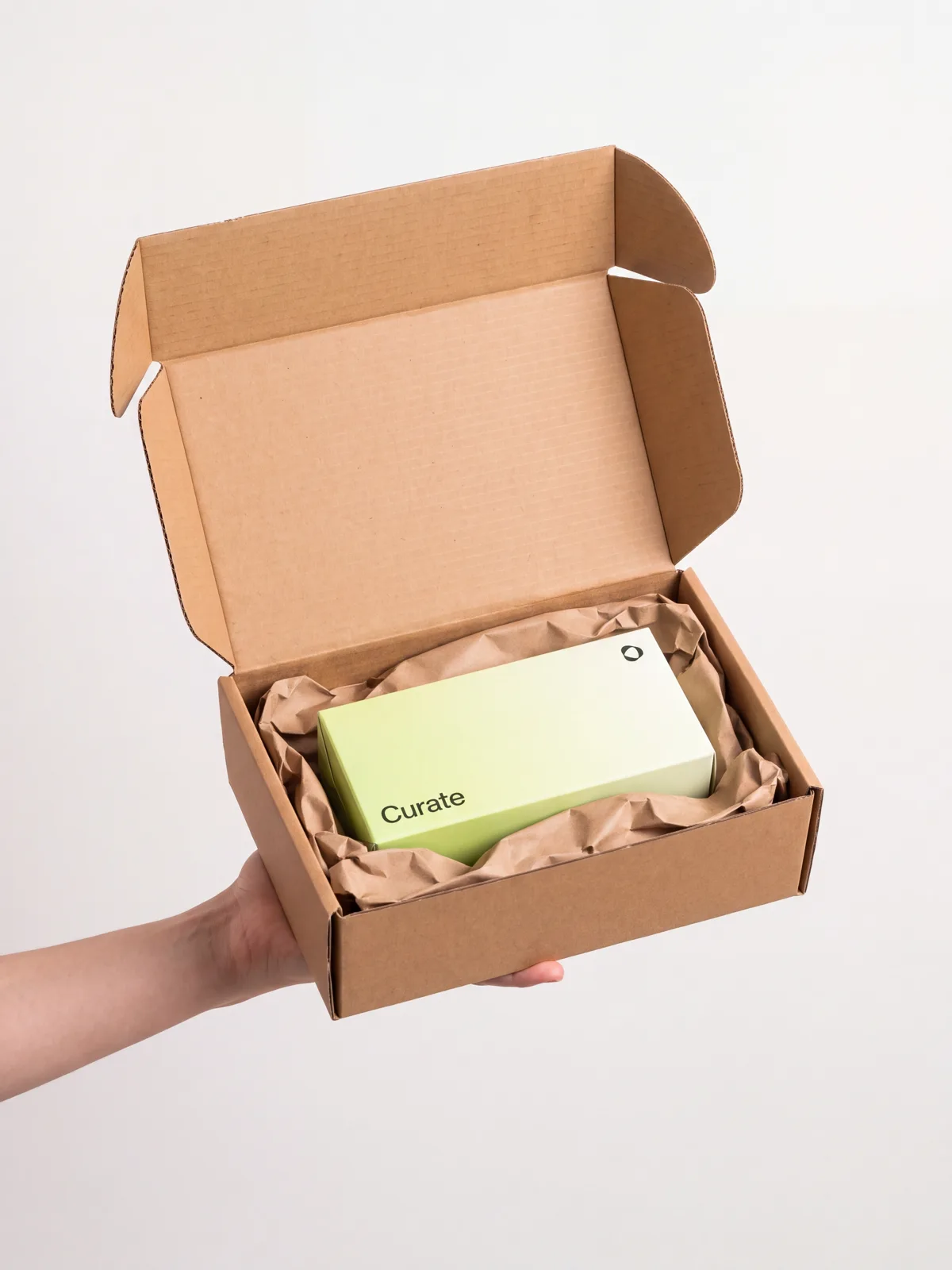 A hand holding an open, unmarked cardboard delivery box with the Curate carton inside on kraft paper